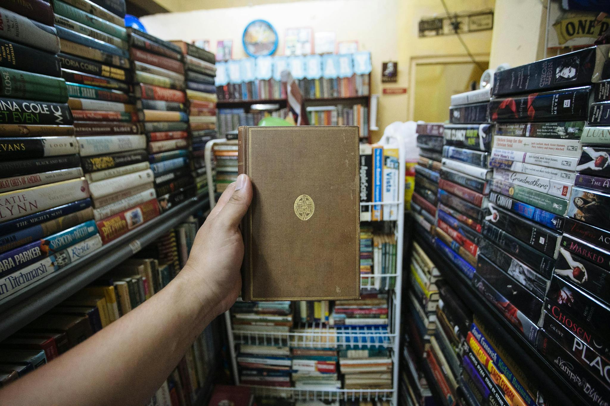 Person holding a vintage hardcover book in a crowded bookstore filled with shelves of various books.